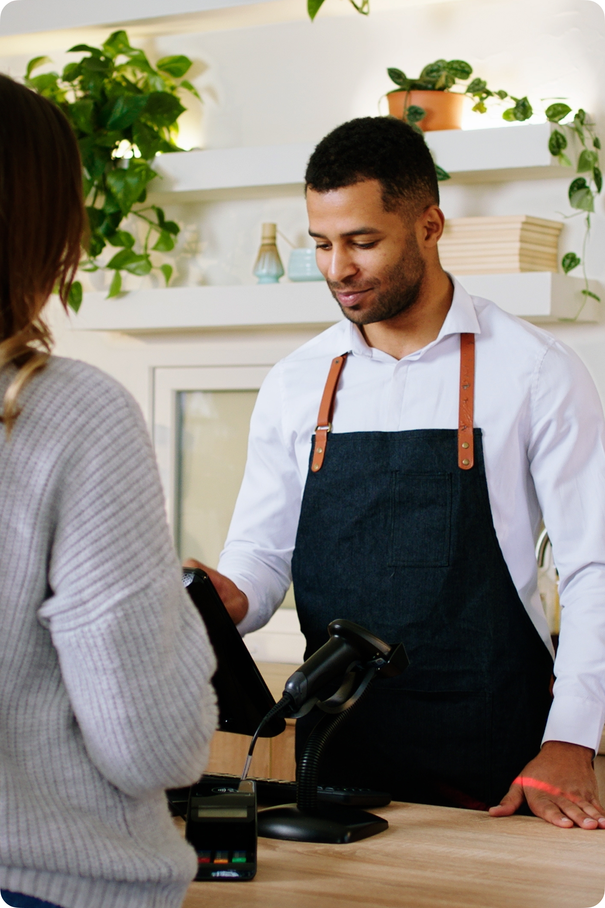 A customer stands at a checkout counter while a store employee wearing a dark apron presents a payment terminal.